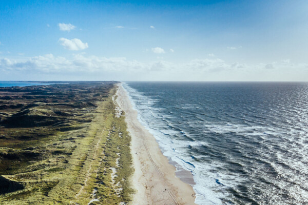 Die Küste der Insel Sylt von oben fotografiert. Es ist auf der linken Seite eine grüne Dünenlandschaft und auf der rechten Seite ein breiter Sandstrand und die Nordsee zu sehen.