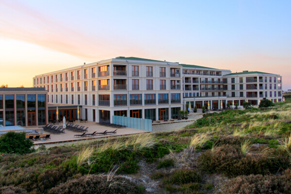 Das Bild zeigt eine Luftaufnahme eines großen, modernen Hotels auf Sylt am Strand. Das Hotel ist von Dünenlandschaften umgeben.  Im Vordergrund führt eine Treppe durch die Dünen, und im Hintergrund ist das ruhige Meer zu sehen. Das klare Wetter und der blaue Himmel verleihen dem Foto eine ruhige, erholsame Atmosphäre.