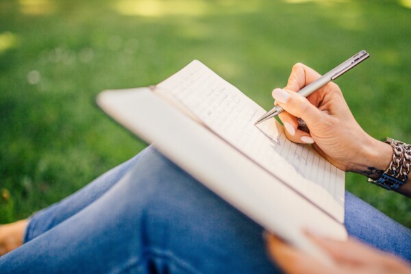 Ein Notizbuch, in welches geschrieben wird, liegt auf einem Schoß. Eine Frauenhand schreibt mit einem silbernen Stift in das Buch. 
