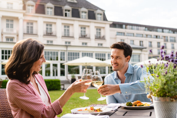 Eine frau und ein Mann stoßen mit Weingläasern beim Essen vor dem A-ROSA Travemünde an.