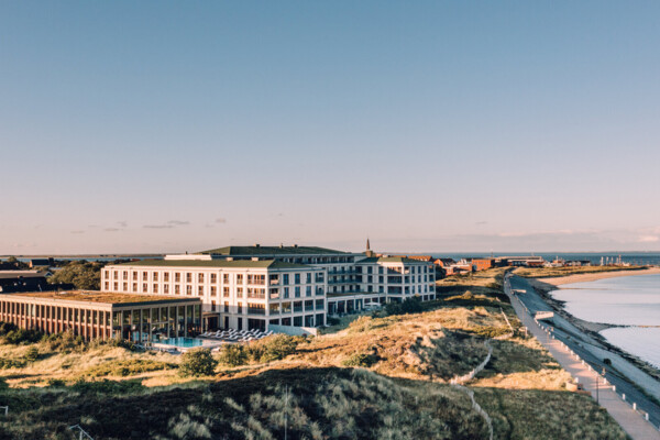Luftaufnahme eines großen, modernen Hotelgebäudes mit mehreren Etagen, eingebettet in Dünenlandschaft mit Blick auf eine Strandpromenade und das Meer im Hintergrund, bei klarem Himmel am Tag.