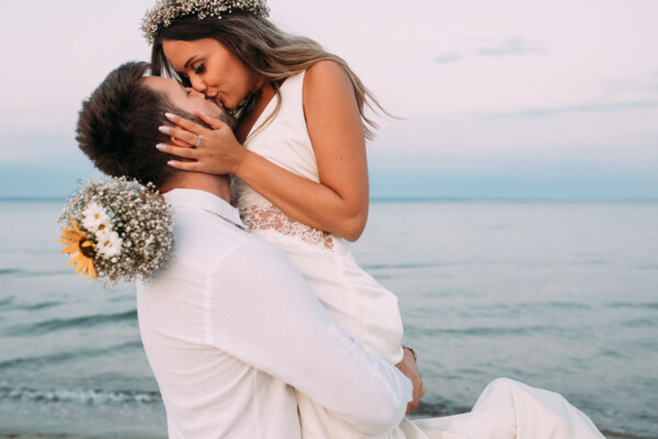 Ein frisch vermähltes Paar umarmt sich liebevoll am Strand. Der Bräutigam in einem weißen Hemd hebt die Braut, die ein weißes Hochzeitskleid und einen Blumenkranz im Haar trägt, hoch. Sie küssen sich zärtlich vor der malerischen Kulisse des ruhigen Meeres bei Dämmerung.