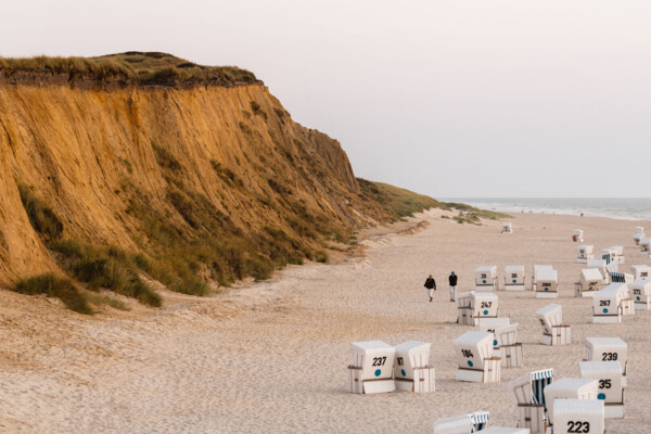 Ein ruhiger Abend an einem sandigen Strand mit einer Klippe im Hintergrund. Im Vordergrund sieht man mehrere weiße Strandkörbe, die typisch für die deutsche Nordseeküste sind. Ein paar Spaziergänger genießen die friedliche Szenerie in der Dämmerung, während das sanfte Meer im Hintergrund die Szene abrundet