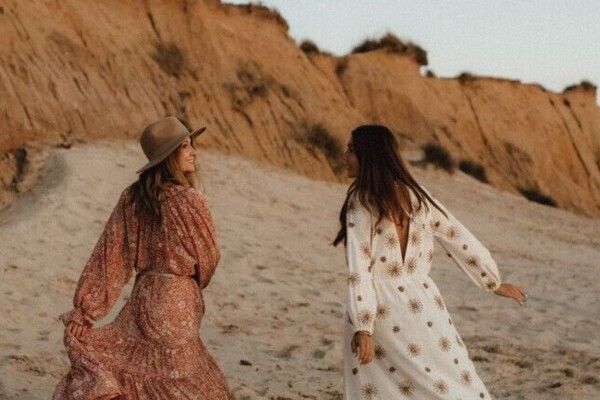 Zwei Frauen in langen Sommerkleidern laufen glücklich entlang eines Strandes, welcher durch die untergehende Sonne in warmes Licht getaucht ist. 
