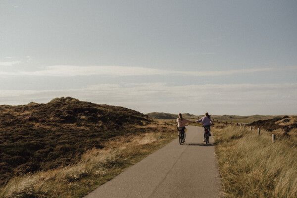 Zwei Frauen fahren auf Fahrrädern, handhaltend, auf einem Asphaltweg durch die Dünen. 