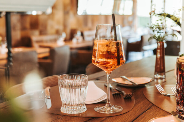 Stilvoll gedeckter Tisch im Restaurant Kaps mit einem Glas Roséwein im Vordergrund, das im warmen Licht des Interieurs glänzt. Im Hintergrund eine gemütliche und einladende Atmosphäre mit gedämpfter Beleuchtung und modernem, alpinem Dekor.