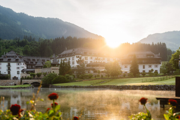 Malerischer Blick auf das A-ROSA Hotels in Kitzbühel, Österreich, bei Sonnenaufgang. Das Hotel zeigt traditionelle alpine Architektur mit weißen Wänden und dunklen Holzbalkonen. Im Vordergrund spiegelt sich das aufgehende Sonnenlicht im ruhigen Wasser eines Sees, umrahmt von blühenden roten Rosen und einer Skulptur am Ufer. Im Hintergrund erheben sich grüne, waldige Hügel, die das idyllische Setting abrunden.