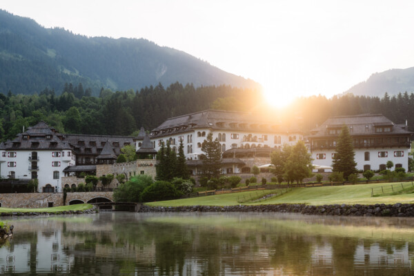 Ein großes, traditionelles Gebäude am Ufer eines Sees, umgeben von bewaldeten Hügeln, mit der aufgehenden Sonne im Hintergrund