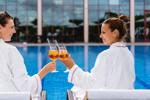 Zwei Frauen stoßen im Bademantel am Pool mit einem Getränk an.